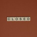 Minimalist image with Scrabble tiles spelling 'CLOSED' on a brown surface.