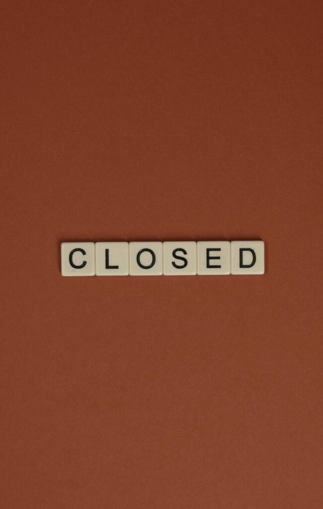 Minimalist image with Scrabble tiles spelling 'CLOSED' on a brown surface.